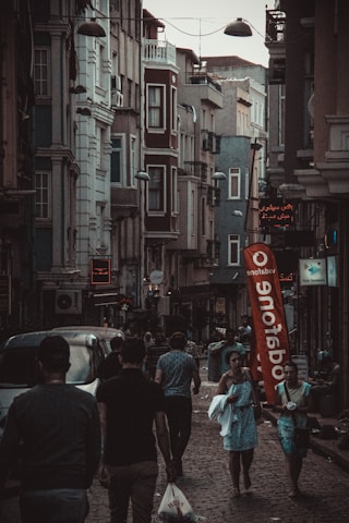 A narrow, bustling street with people walking along sidewalks between tall, closely-packed urban buildings. The architecture features multiple stories with various windows and balconies. Street lights hang overhead, and a large red Vodafone flag stands out in the scene. Several shops have illuminated signs in different languages.