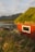 A cozy red cabin with a grass-covered roof is nestled amidst lush greenery, with tall grass and wildflowers in the foreground. The scene is set against a backdrop of calm water and majestic mountains under a partly cloudy sky, capturing the serene beauty of nature.