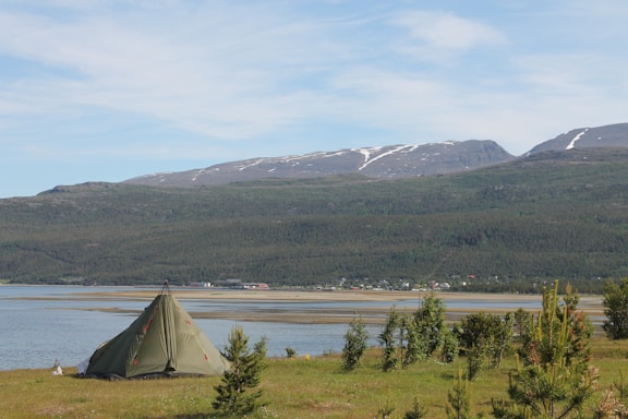 A green tent is pitched on a grassy field near a large body of water, with lush green trees scattered along the shore. In the background, expansive mountains with patches of snow contrast against a blue sky with light clouds. Distant houses or buildings can be observed along the water's edge.