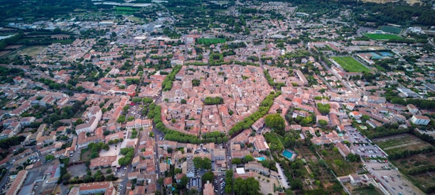 An aerial view of a densely built urban area featuring tightly packed buildings with a mix of residential and commercial structures. A prominent circular arrangement of buildings and roads is visible at the center, surrounded by areas of greenery. Roads intersect the area, leading to and from the central circle. The outskirts show more open spaces with fields and larger structures, possibly indicating industrial or recreational areas.