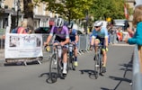 Cyclists are competing in a road race, surrounded by spectators behind metal barriers. The riders wear helmets and colorful cycling gear, and they are focused on the race. A banner for an event planning service is visible on the left. Trees and buildings line the background under a clear sky.