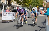 Cyclists are competing in a road race, surrounded by spectators behind metal barriers. The riders wear helmets and colorful cycling gear, and they are focused on the race. A banner for an event planning service is visible on the left. Trees and buildings line the background under a clear sky.