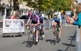 Cyclists are competing in a road race, surrounded by spectators behind metal barriers. The riders wear helmets and colorful cycling gear, and they are focused on the race. A banner for an event planning service is visible on the left. Trees and buildings line the background under a clear sky.