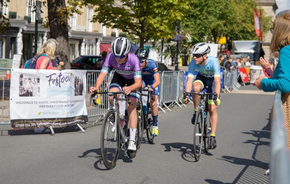 Cyclists are competing in a road race, surrounded by spectators behind metal barriers. The riders wear helmets and colorful cycling gear, and they are focused on the race. A banner for an event planning service is visible on the left. Trees and buildings line the background under a clear sky.