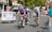 Cyclists are competing in a road race, surrounded by spectators behind metal barriers. The riders wear helmets and colorful cycling gear, and they are focused on the race. A banner for an event planning service is visible on the left. Trees and buildings line the background under a clear sky.