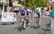 Cyclists are competing in a road race, surrounded by spectators behind metal barriers. The riders wear helmets and colorful cycling gear, and they are focused on the race. A banner for an event planning service is visible on the left. Trees and buildings line the background under a clear sky.