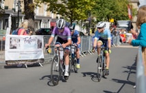 Cyclists are competing in a road race, surrounded by spectators behind metal barriers. The riders wear helmets and colorful cycling gear, and they are focused on the race. A banner for an event planning service is visible on the left. Trees and buildings line the background under a clear sky.