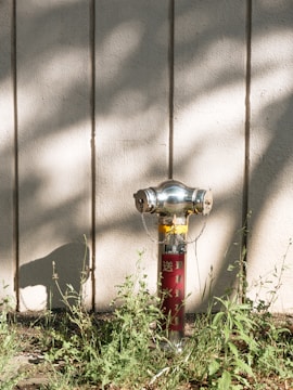 A fire hydrant stands against a textured wall with vertical lines. The hydrant is metallic with yellow and red components and has chains attached to its sides. Surrounding the base are patches of green grass and weeds. Shadows from nearby trees fall across the wall and ground, adding a dappled light effect.