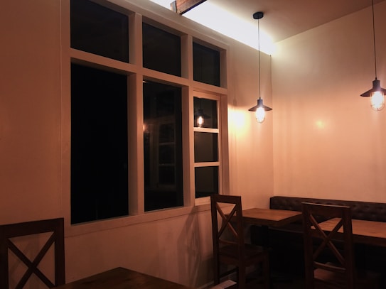 A cozy indoor dining area with wooden tables and chairs. Soft, warm lighting hangs from the ceiling, creating a peaceful ambiance. The corner features a tufted bench seat against the wall, and a large window panel adds to the decor.