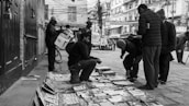 A busy street scene features several people gathered around a small market stall selling printed materials laid out on the ground. One person is seated on a stool, engaging with customers, while others browse newspapers and other publications. The urban setting includes buildings with signage and tangled overhead wires, conveying an everyday street life atmosphere.