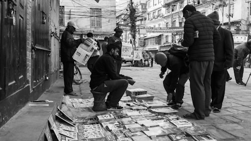 A busy street scene features several people gathered around a small market stall selling printed materials laid out on the ground. One person is seated on a stool, engaging with customers, while others browse newspapers and other publications. The urban setting includes buildings with signage and tangled overhead wires, conveying an everyday street life atmosphere.