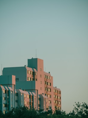 A modern apartment building with multiple floors is seen against a clear sky. The structure is bathed in soft sunlight, casting warm tones across its facade. The design features rectangular windows, and the composition conveys a sense of tranquility. At the base, trees add a natural element to the urban setting.