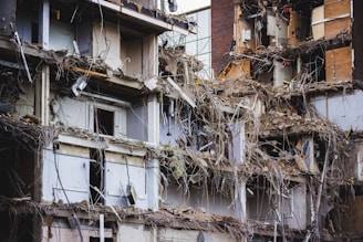A heavily damaged and partially collapsed building with exposed interiors and debris scattered throughout. The structure appears old, with visible steel bars, electrical wiring, and shattered walls. The scene suggests a recent demolition or natural disaster aftermath.