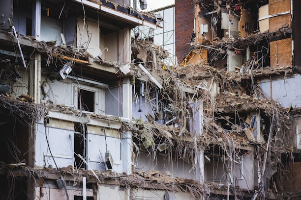 A heavily damaged and partially collapsed building with exposed interiors and debris scattered throughout. The structure appears old, with visible steel bars, electrical wiring, and shattered walls. The scene suggests a recent demolition or natural disaster aftermath.