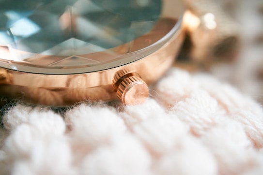 A close-up of a rose gold watch resting on a soft, textured, cream-colored fabric. The watch features a faceted, crystal-like surface and a prominent crown for adjusting time.