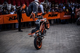 A person is performing a stunt on a motorcycle, riding on the front wheel while being airborne. The motorcycle has vibrant orange accents and is positioned in front of a crowd. The audience is gathered behind safety barriers adorned with orange KTM branding.