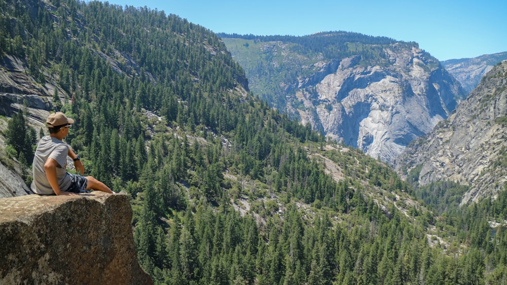 Hombre con camisa gris de cuello redondo sentado en el paisaje de la montaña del borde de la roca