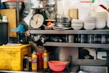 A cluttered kitchen scene features stacks of white bowls and metal bowls on a shelf. There is a scale showing a dial, various condiment bottles, a yellow container, and kitchen utensils scattered around. Plastic cups, a chopping board, and other kitchen equipment are also visible.