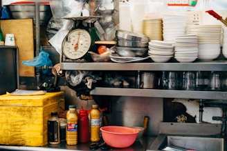 A cluttered kitchen scene features stacks of white bowls and metal bowls on a shelf. There is a scale showing a dial, various condiment bottles, a yellow container, and kitchen utensils scattered around. Plastic cups, a chopping board, and other kitchen equipment are also visible.