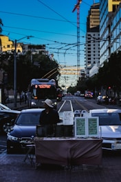A city street scene featuring a vendor standing behind a table with jewelry displays and a sign reading 'Name Wire.' The vendor is wearing a hat and appears to be waiting for customers. Surrounding the scene are cars and a tall building with a crane, under a clear blue sky. Electrical wires and a bus are visible in the background.