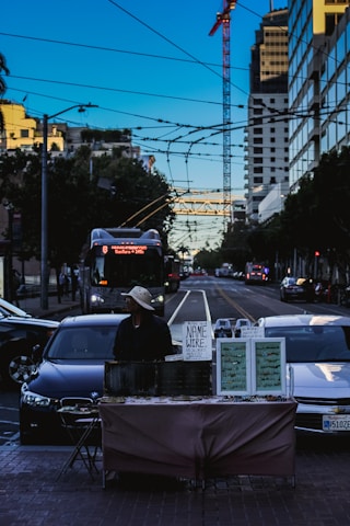 A city street scene featuring a vendor standing behind a table with jewelry displays and a sign reading 'Name Wire.' The vendor is wearing a hat and appears to be waiting for customers. Surrounding the scene are cars and a tall building with a crane, under a clear blue sky. Electrical wires and a bus are visible in the background.