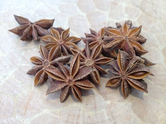 Several star anise pods are arranged on a light wooden surface. The pods have a star-shaped appearance with a dark brown color and a textured, rough surface. The seeds inside the pods are glossy and dark, providing a contrast to the outer shell.