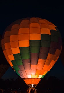 A brightly lit hot air balloon filled with vibrant colors like orange, green, and purple against a dark sky. The flame at the base creates a warm glow that illuminates the balloon's panels. The silhouette of trees can be seen at the bottom.