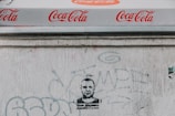 A Coca-Cola branded canopy with repeated logos visible above a textured white wall. The wall has various faded graffiti and a stenciled portrait of a person with text beneath.