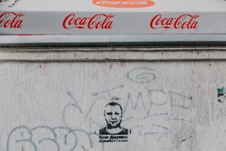 A Coca-Cola branded canopy with repeated logos visible above a textured white wall. The wall has various faded graffiti and a stenciled portrait of a person with text beneath.