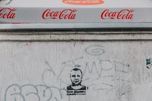 A Coca-Cola branded canopy with repeated logos visible above a textured white wall. The wall has various faded graffiti and a stenciled portrait of a person with text beneath.