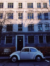 A classic white Volkswagen Beetle is parked on a quiet street in front of a large, traditional European-style building adorned with windows featuring green frames. Shadows of bare tree branches are cast onto the building facade. A bicycle with an orange frame is partially visible behind the car.