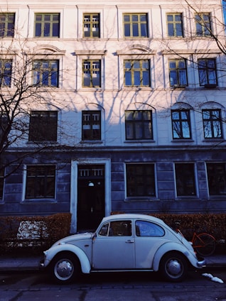 A classic white Volkswagen Beetle is parked on a quiet street in front of a large, traditional European-style building adorned with windows featuring green frames. Shadows of bare tree branches are cast onto the building facade. A bicycle with an orange frame is partially visible behind the car.