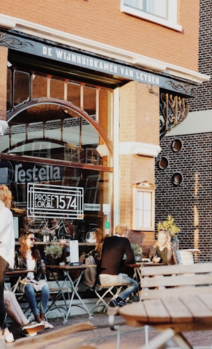 A small group of people is seated at outdoor tables of a caf&eacute; or restaurant on a sunny day. The establishment features large glass windows with signage indicating 'Proef Lokaal 1574' and 'Geopend'. The building has a vintage brick facade with decorative elements. Customers appear relaxed and engaged in conversation or using their phones.