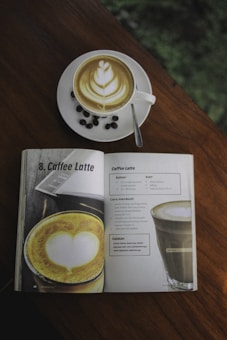 A coffee cup with latte art sits on a wooden table alongside a book that displays a recipe for a caffe latte. Coffee beans are scattered around the saucer.