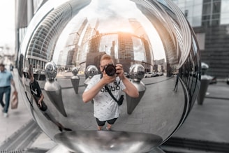 A person holding a camera is reflected in a large, shiny metallic sphere. Surrounding the sphere are buildings of a cityscape with modern architecture. The reflection also includes several smaller metallic spheres and a street scene with pedestrians and vehicles.