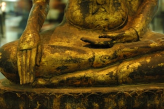 A close-up view of a golden Buddha statue, focusing on the intricately carved hands and torso. The surface displays wear, revealing a textured patina that adds depth to the piece. The position of the hands suggests a meditative pose.