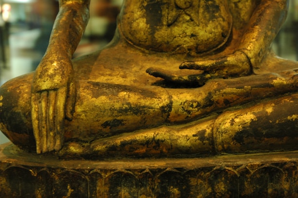 A close-up view of a golden Buddha statue, focusing on the intricately carved hands and torso. The surface displays wear, revealing a textured patina that adds depth to the piece. The position of the hands suggests a meditative pose.