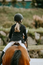 A person wearing a helmet and riding attire is seen from behind while riding a brown horse. The setting appears to be outdoors with blurred greenery in the background. The rider has long hair tied back, and the horse is positioned squarely in the center, showing its tail prominently.
