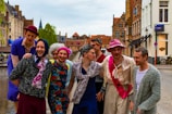A group of people dressed in colorful, eccentric clothing pose happily in a historic European city setting. They are wearing a mix of hats, wigs, and vibrant clothing, with one person wearing a pink sash. The background includes traditional brick buildings and a canal, suggesting a charming old-town atmosphere.