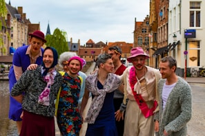 A group of people dressed in colorful, eccentric clothing pose happily in a historic European city setting. They are wearing a mix of hats, wigs, and vibrant clothing, with one person wearing a pink sash. The background includes traditional brick buildings and a canal, suggesting a charming old-town atmosphere.