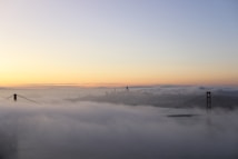 A scenic view captures the Golden Gate Bridge partially enveloped in fog during a tranquil sunrise or sunset. The skyline of a city is faintly visible through the fog, set against a softly lit sky transitioning from pale blue to warm orange tones.