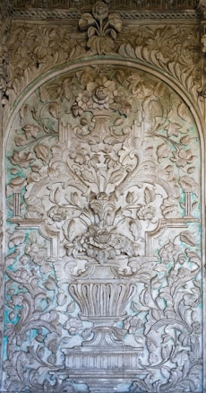 An intricate stone relief features elaborate floral and leaf patterns carved in a symmetrical design. The centerpiece displays a large flower vase with a bouquet of flowers and leaves extending outward. The background reveals subtle hints of greenish patina, adding depth and contrast.