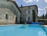 A quaint stone house with wooden shutters and ivy growing on the walls is situated next to a clear, blue swimming pool. The pool has an inflatable swan and a rolled-up pool cover on the side. The sky is clear with some fluffy white clouds, and the scene is bordered by greenery.