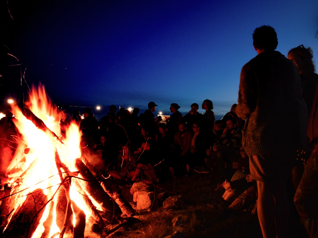 people gather near bonfire during night, A warming bonfire after the sun goes down draws the festival crowd in.<p></figure>I was very taken with the glow of the fire creating a contrast with the darkening sky and at the same time some back light that created some attractive silhouettes.</p><p>The flame blur and trails from fire embers shooting up in the heat added a pleasing dynamic feel to the shot that contrasts with the relaxed onlookers.