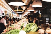 A busy indoor market with a variety of fresh produce displayed on stalls. Customers and vendors are interacting amid vibrant arrays of vegetables and fruits. Overhead, a series of industrial lights cast a warm glow, enhancing the lively atmosphere. Signs and boxes add to the bustling, crowded setting.