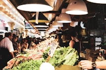 A busy indoor market with a variety of fresh produce displayed on stalls. Customers and vendors are interacting amid vibrant arrays of vegetables and fruits. Overhead, a series of industrial lights cast a warm glow, enhancing the lively atmosphere. Signs and boxes add to the bustling, crowded setting.
