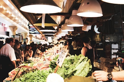 A busy indoor market with a variety of fresh produce displayed on stalls. Customers and vendors are interacting amid vibrant arrays of vegetables and fruits. Overhead, a series of industrial lights cast a warm glow, enhancing the lively atmosphere. Signs and boxes add to the bustling, crowded setting.