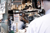 A market scene with a vendor wearing a cap and sunglasses behind a counter, engaging with a customer. Signs advertise fresh Alaskan sockeye salmon priced at 11.99 per pound. Various dried fish hang above, and shelves filled with jars and bottles of spices and condiments are in the background. The area is lively with multiple signs and price tags, giving a bustling feel.