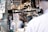 A market scene with a vendor wearing a cap and sunglasses behind a counter, engaging with a customer. Signs advertise fresh Alaskan sockeye salmon priced at 11.99 per pound. Various dried fish hang above, and shelves filled with jars and bottles of spices and condiments are in the background. The area is lively with multiple signs and price tags, giving a bustling feel.