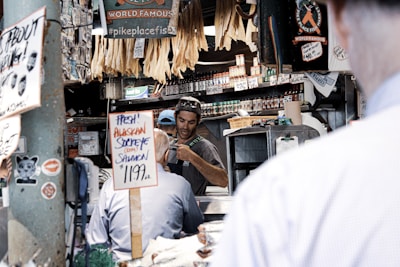 A market scene with a vendor wearing a cap and sunglasses behind a counter, engaging with a customer. Signs advertise fresh Alaskan sockeye salmon priced at 11.99 per pound. Various dried fish hang above, and shelves filled with jars and bottles of spices and condiments are in the background. The area is lively with multiple signs and price tags, giving a bustling feel.